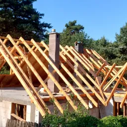 Wznoszenie drewnianej więźby dachowej na murowanym domu jednorodzinnym, widoczne kominy, częściowo pokryte płyty OSB, tło stanowią drzewa iglaste i błękitne niebo.