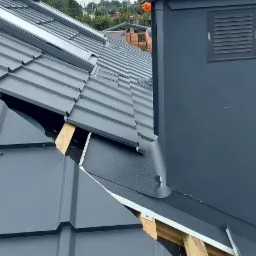 Montaż szarej blachy na dachu z widoczną konstrukcją drewnianą, kominem i fragmentem sylwetki dekarza w tle pochmurnego nieba.