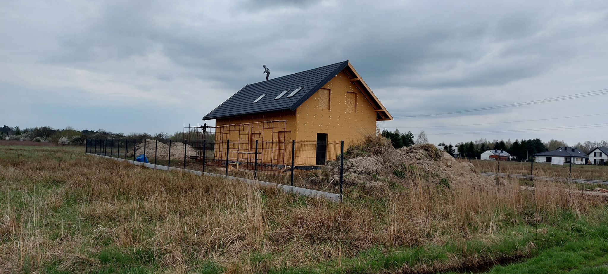 Dom w trakcie budowy z czarnym dachem i dwoma oknami dachowymi, widoczny pracownik na dachu, elewacja w trakcie prac, rusztowania przy ścianie, otoczenie: trawa, ziemia, ogrodzenie, pochmurne niebo.