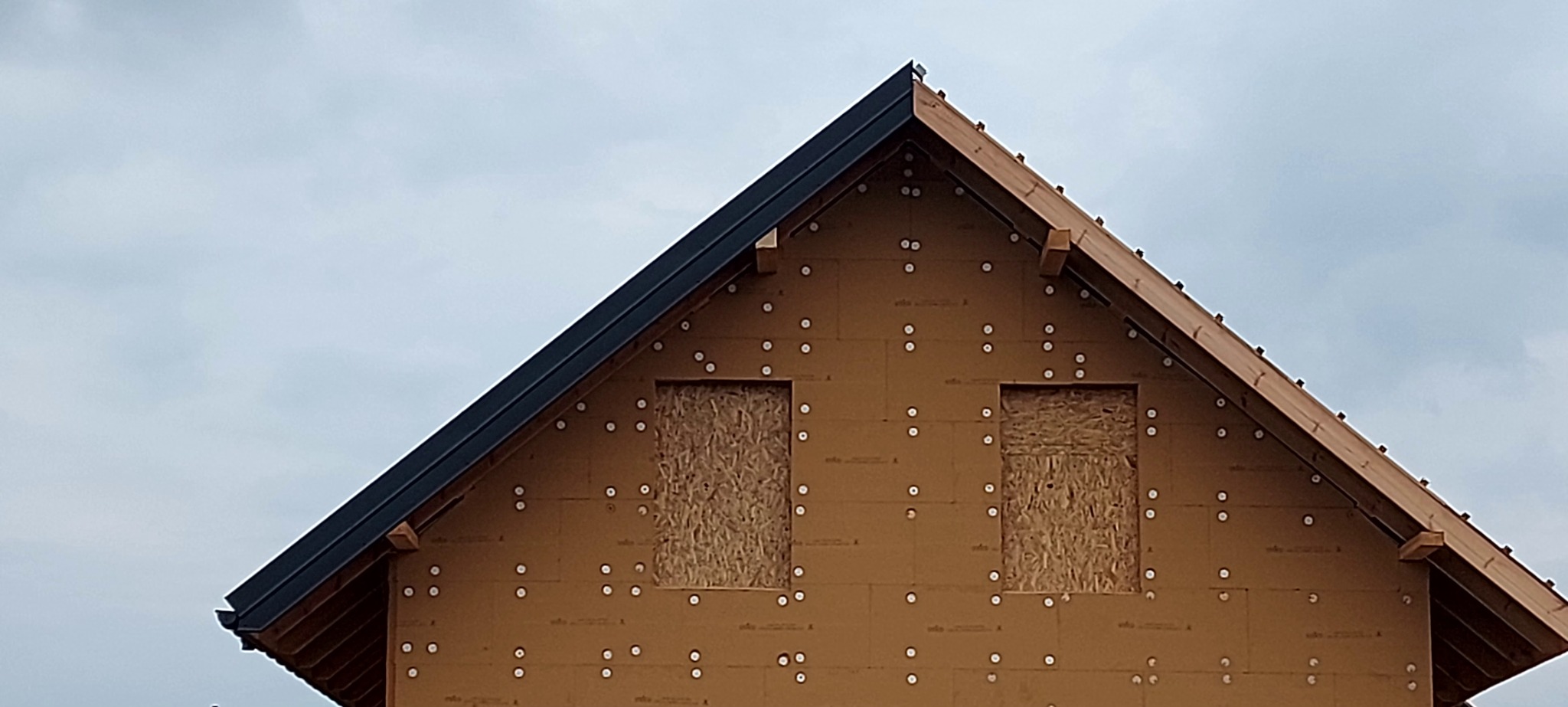 Szczyt budynku w trakcie budowy z widoczną izolacją zewnętrzną, deskami zakrywającymi otwory okienne oraz zamontowanymi obróbkami blacharskimi w kolorze grafitowym.