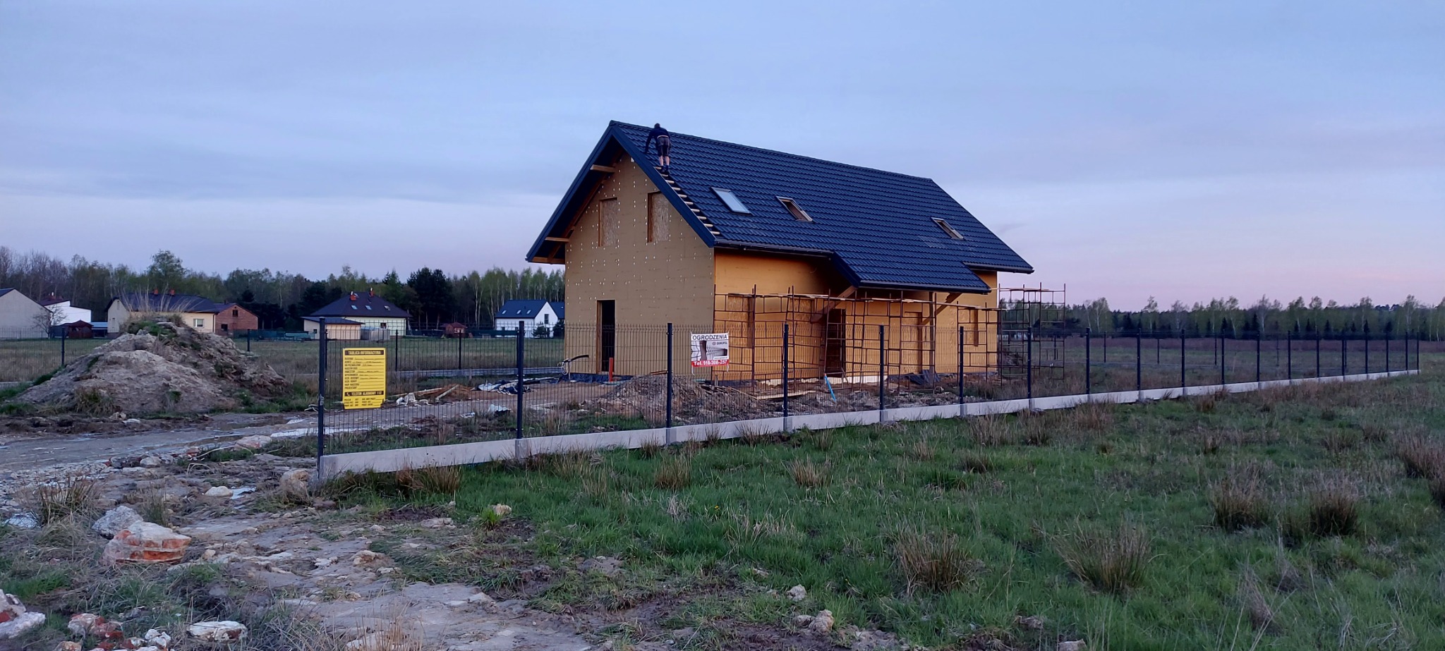 Budowa domu jednorodzinnego w trakcie, widoczna konstrukcja dachu z oknami dachowymi, ściany obłożone płytami izolacyjnymi, częściowo zamontowane rusztowania, w tle inne domy i drzewa, ogrodzenie...