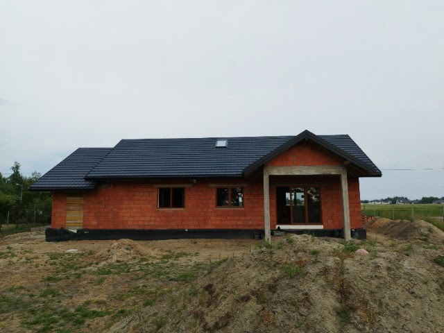 Niedokończony dom jednorodzinny z ciemną dachówką, widoczne cegły, okna, fragment dachu z lukarną i teren budowy z ziemią.