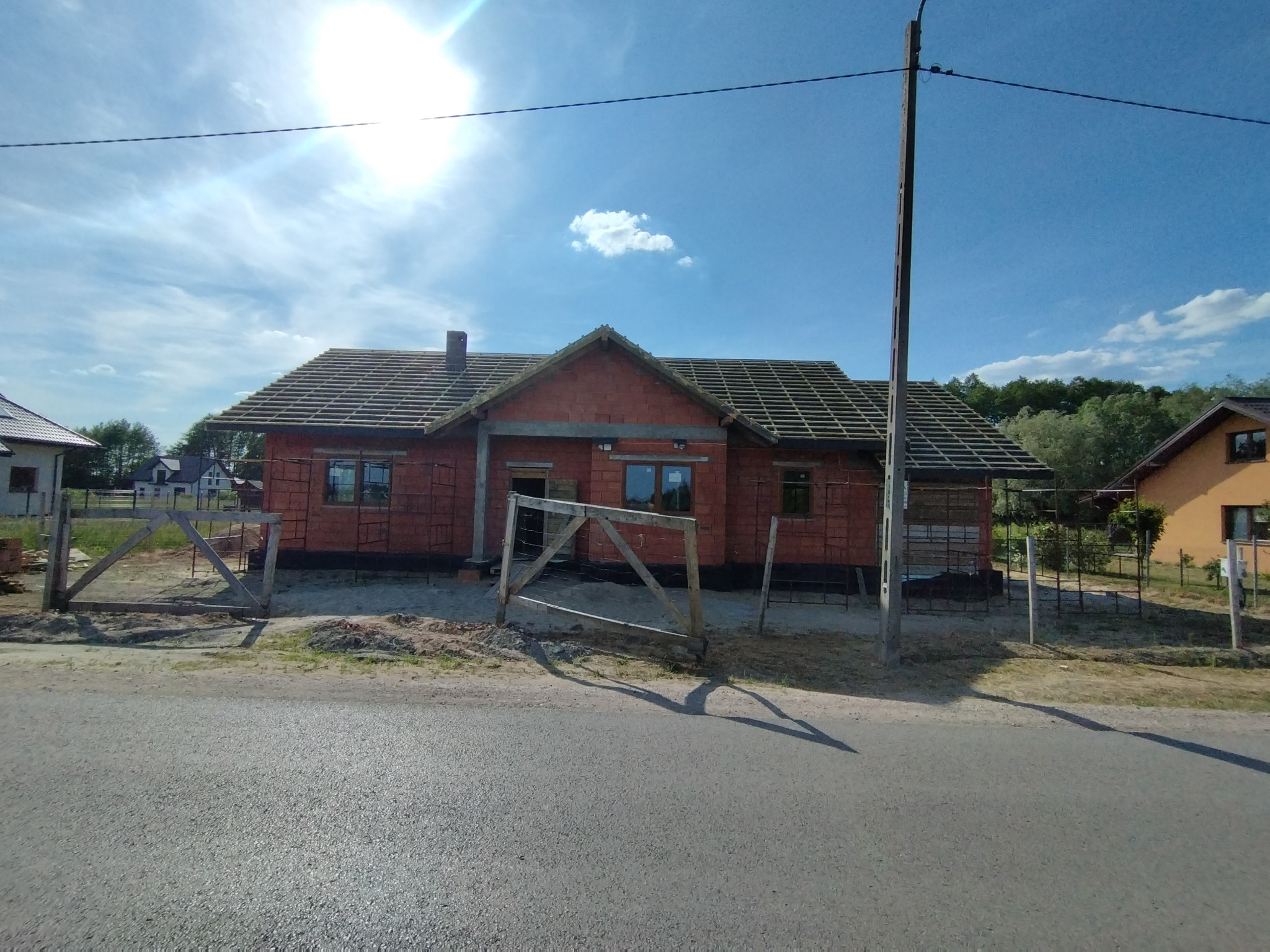 Surowy budynek z czerwonej cegły w trakcie budowy dachu, widoczna więźba dachowa i wstępne pokrycie, otoczony drewnianym ogrodzeniem i słupem energetycznym, na tle błękitnego nieba.