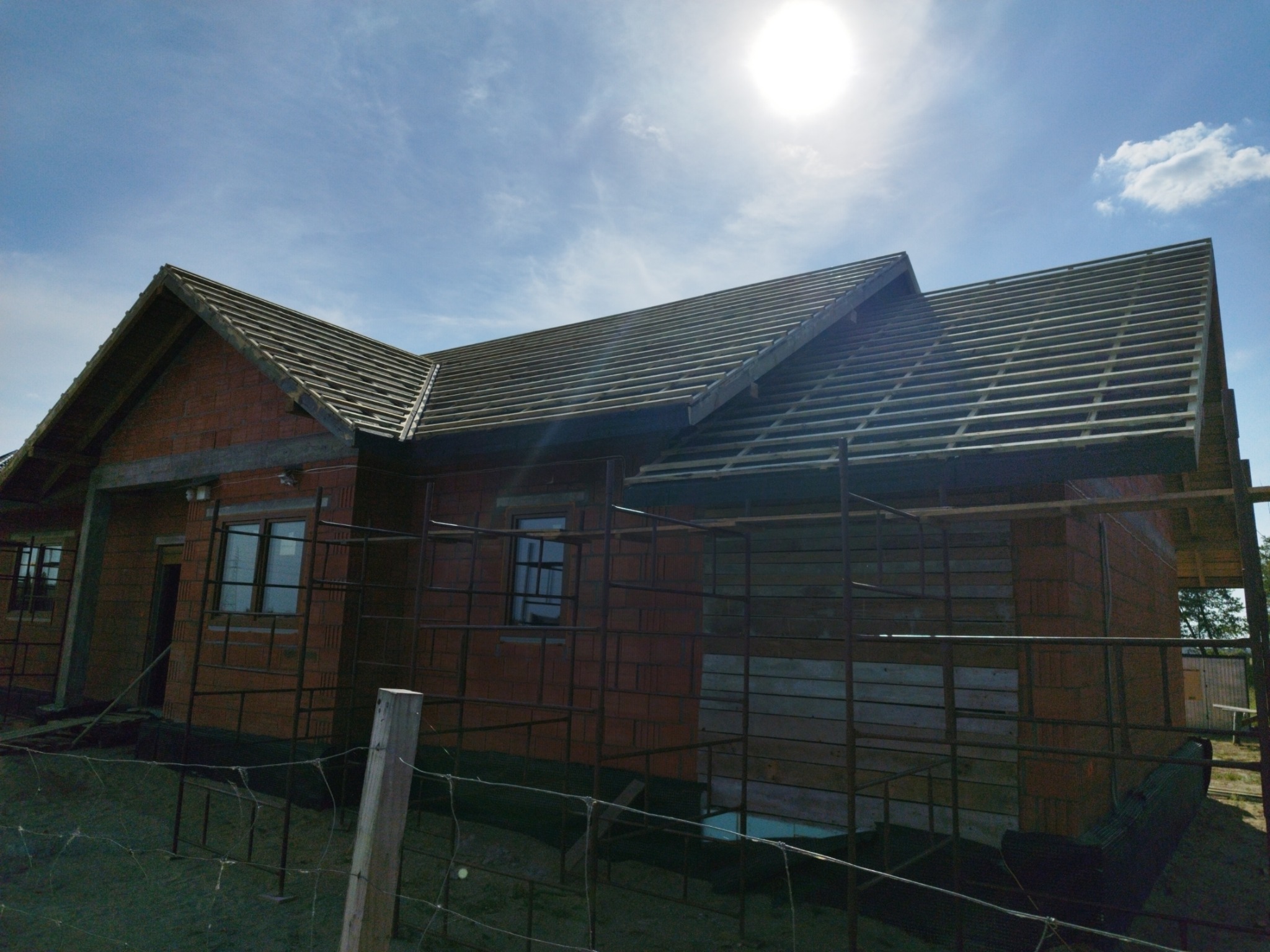 Dom w trakcie budowy z widoczną konstrukcją więźby dachowej, częściowo pokrytą dachówką, w otoczeniu rusztowań i ogrodzenia.