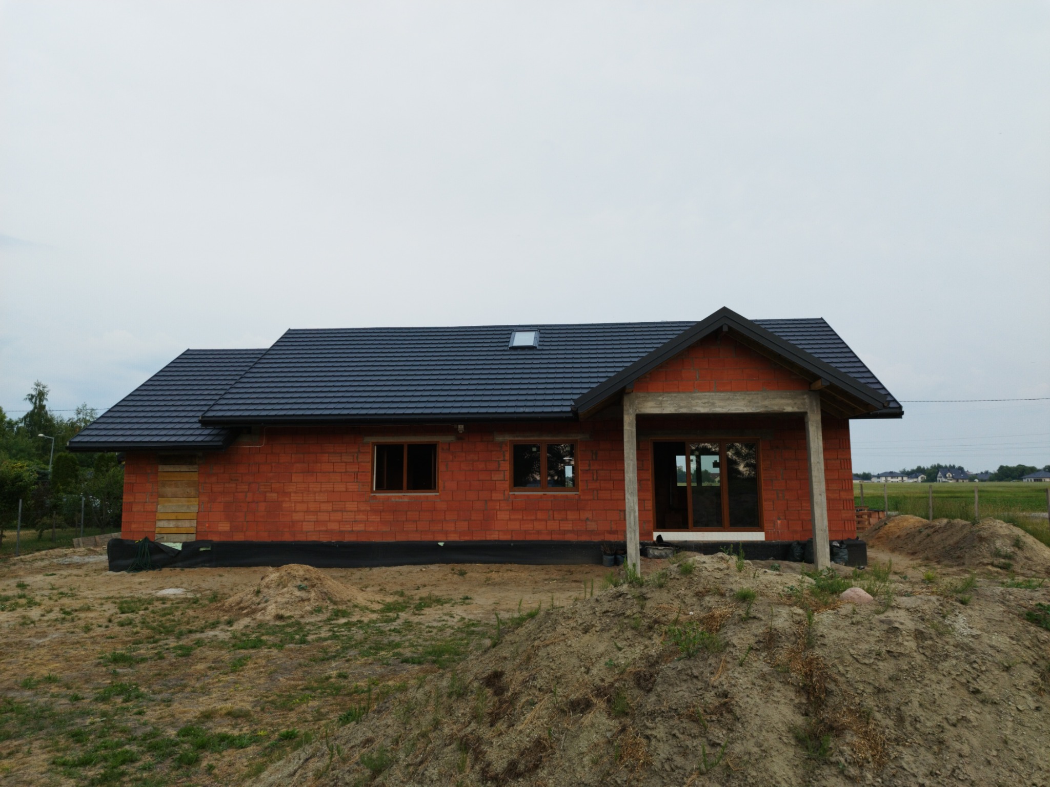 Dom w trakcie budowy z ciemną dachówką, widoczne okna z brązowymi ramami i wejście z drewnianą konstrukcją, otoczony ziemią i trawą.