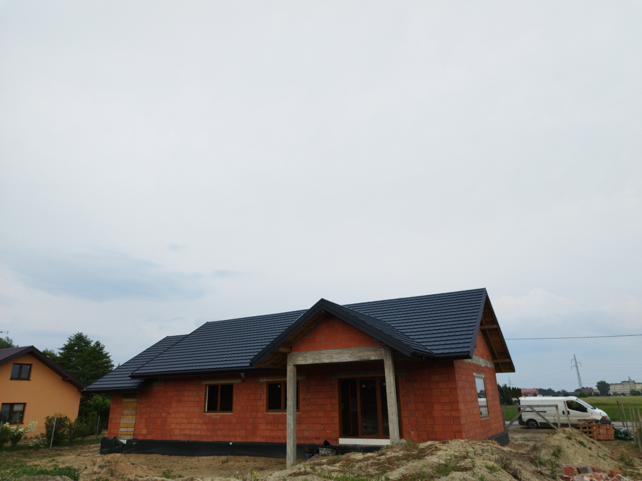 Nowo wybudowany dom z ciemną dachówką w Żabiej Woli, widoczne surowe ściany z cegły i niezadaszone wejście, na tle zachmurzone niebo.