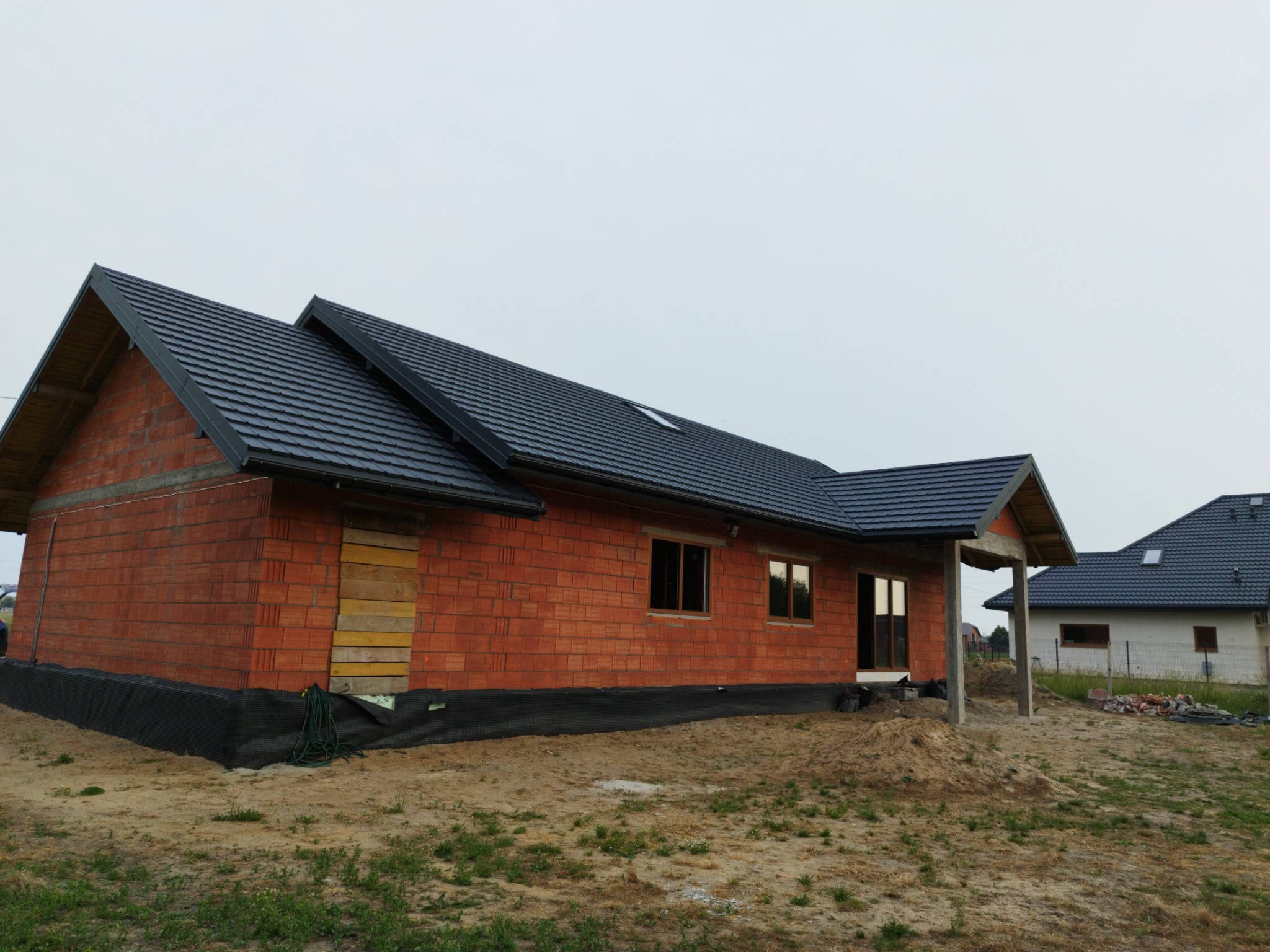 Surowy budynek z czerwonej cegły z ciemnoszarą dachówką, widoczna membrana na fundamencie i tymczasowe zabezpieczenie drzwi deskami, drugi budynek w tle.