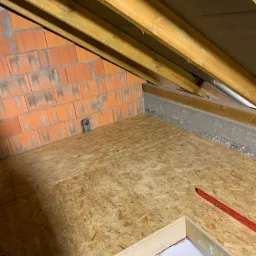 Poddasze w trakcie adaptacji: widoczne drewniane belki konstrukcyjne, ceglana ściana z szarą rurą, płyta OSB na podłodze oraz poziomica w rogu.