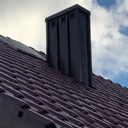 Ciemnoczerwona dachówka ceramiczna z nowoczesnym, ciemnym kominem z metalową obróbką, widok z dołu na tle pochmurnego nieba.