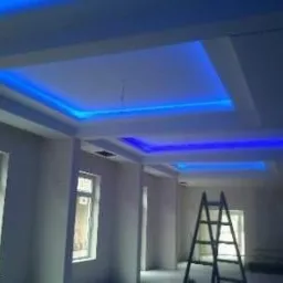 Nowoczesny sufit wielopoziomowy z oświetleniem LED w kolorze niebieskim, widoczna drabina w pustym, jasnym pomieszczeniu w trakcie wykończenia.