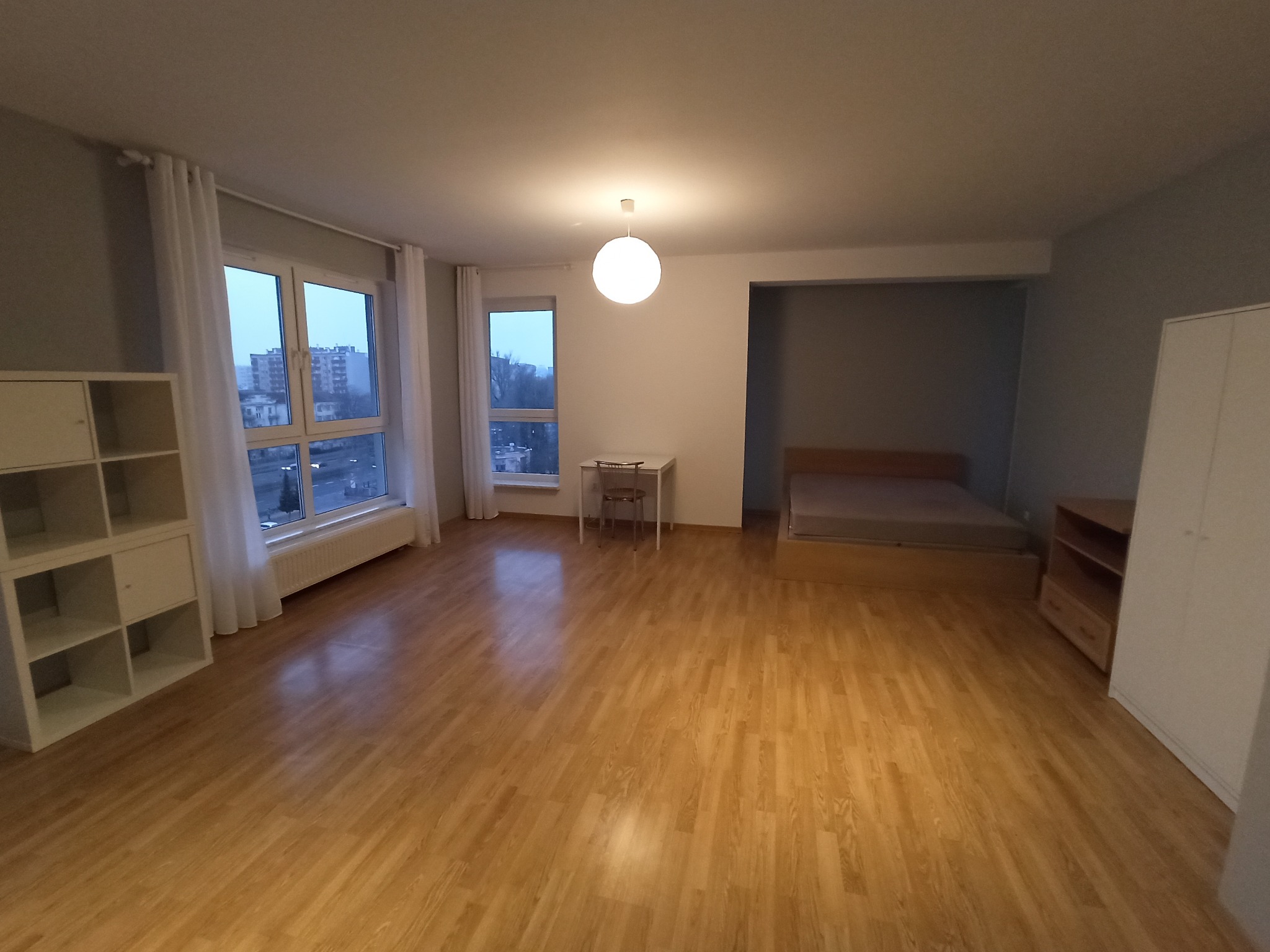 Puste, odświeżone wnętrze kawalerki z drewnianą podłogą, białymi meblami i widokiem na miasto przez okno.