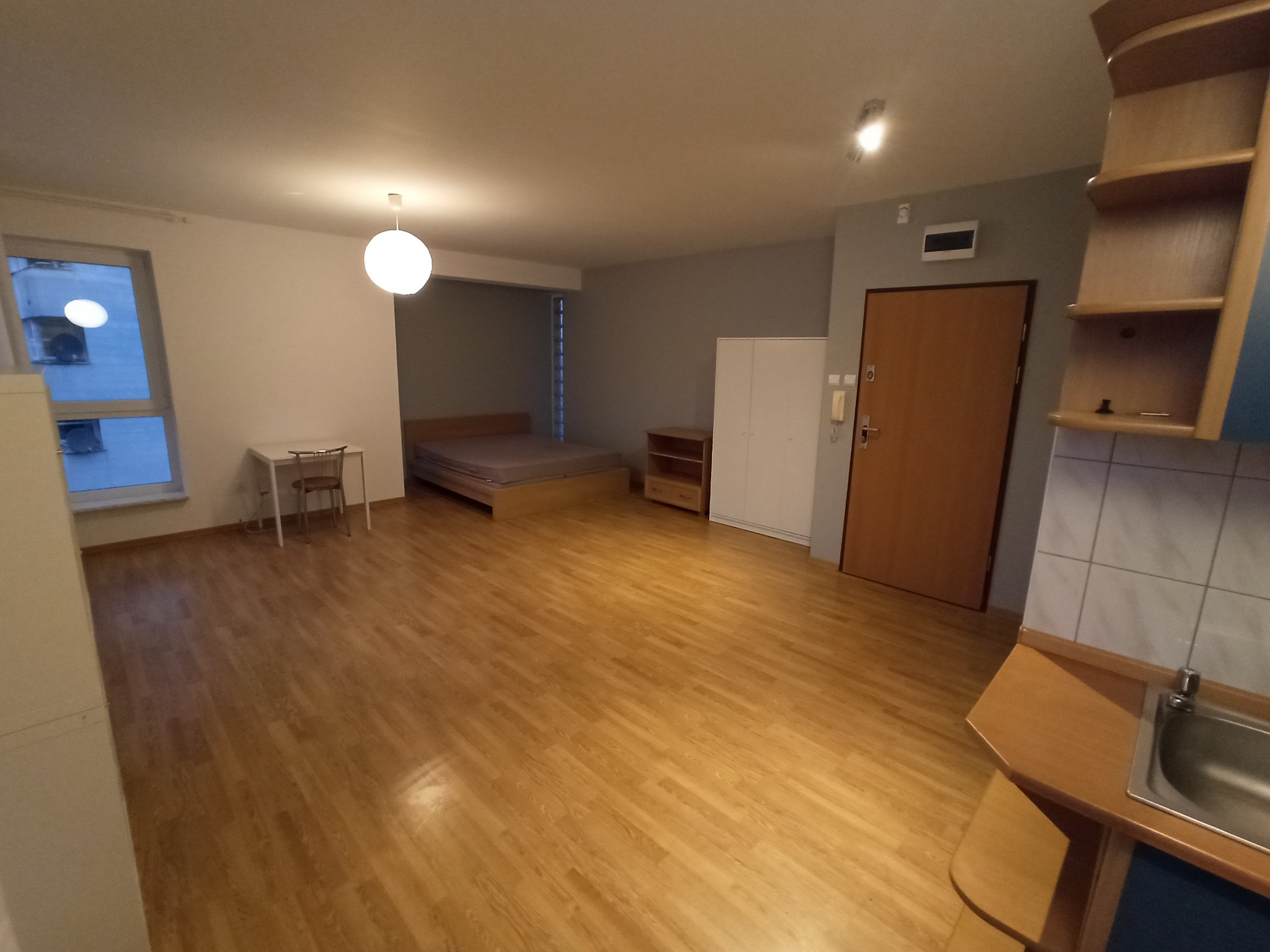 Widok wnętrza wyremontowanej kawalerki z drewnianą podłogą, białymi ścianami, aneksem kuchennym po prawej stronie i łóżkiem w głębi pomieszczenia.