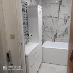 Nowoczesna łazienka z białą zabudową meblową, grzejnikiem drabinkowym i wanną, wykończona płytkami imitującymi marmur.