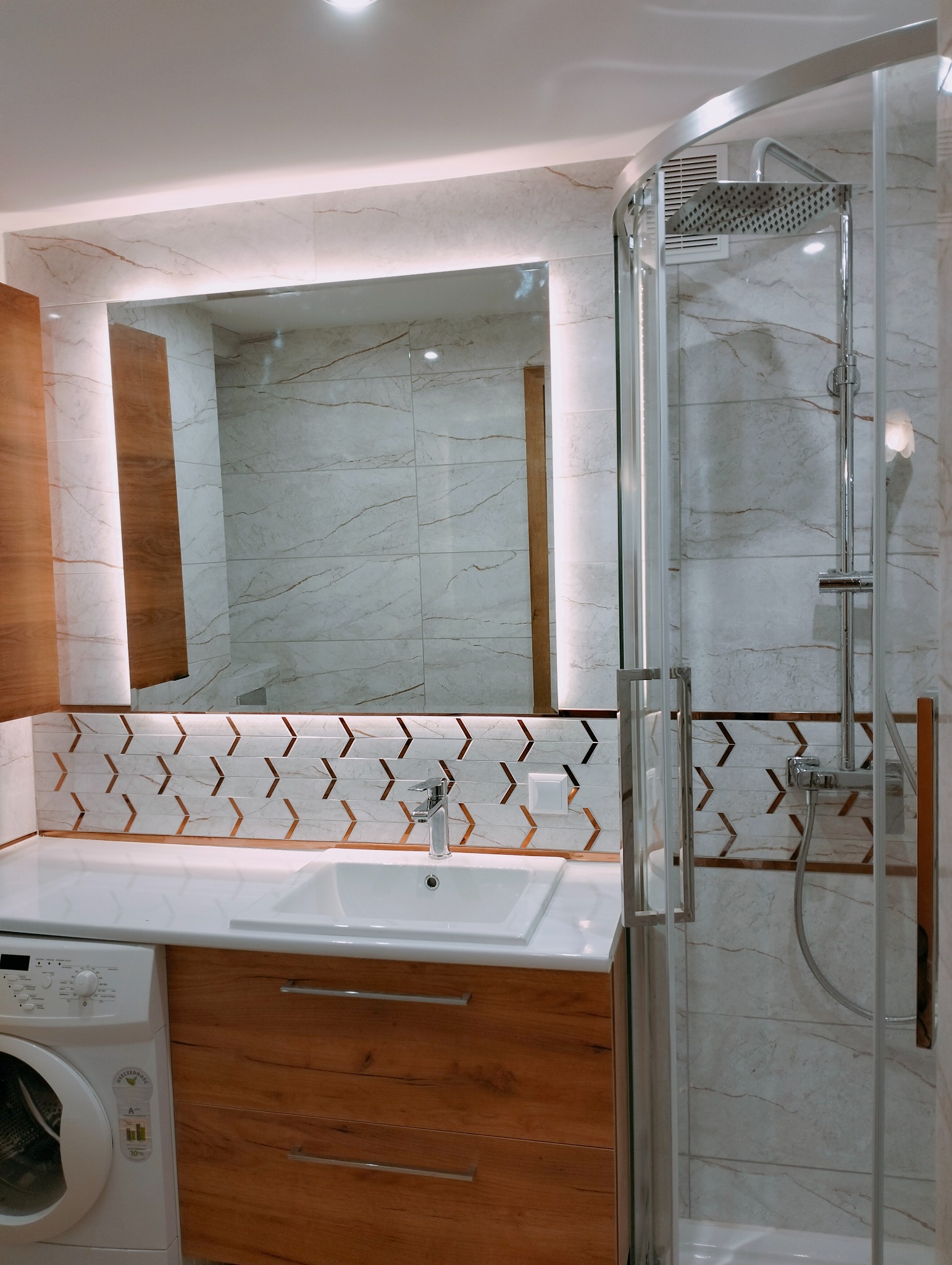 Nowoczesna łazienka z kabiną prysznicową, umywalką wpuszczaną w blat z szafką z drewnopodobnego materiału, pralką obok, lustrem z podświetleniem LED i dekoracyjną mozaiką w kształcie strzałek.