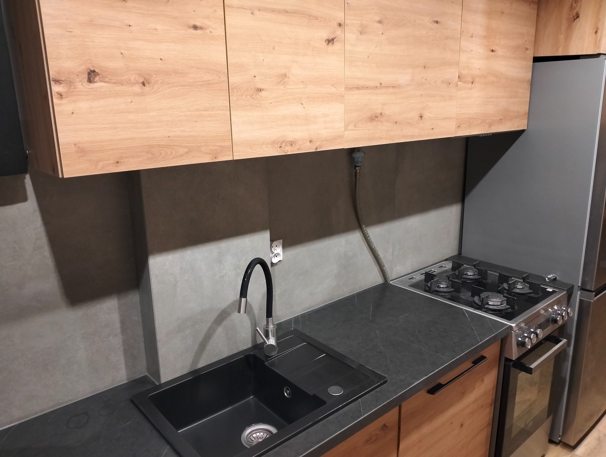 Nowoczesna kuchnia z drewnianymi szafkami, czarnym blatem i zlewozmywakiem, oraz stalową kuchenką gazową i lodówką, widoczny fragment instalacji wodnej.