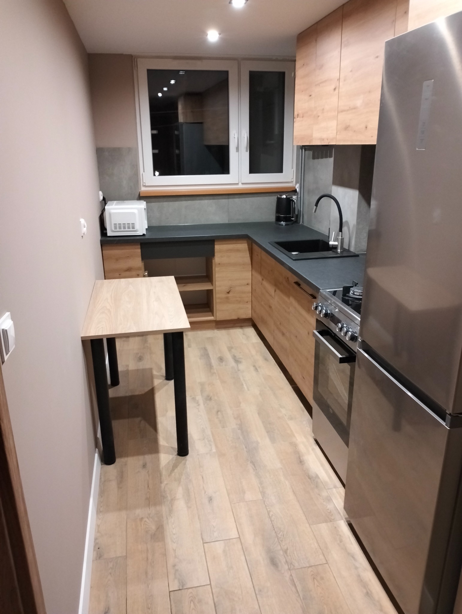 Jasna kuchnia z drewnianymi szafkami, czarnym blatem i stołem z czarnymi nogami, oświetlona punktowo, z widocznym sprzętem AGD, takim jak lodówka i kuchenka.