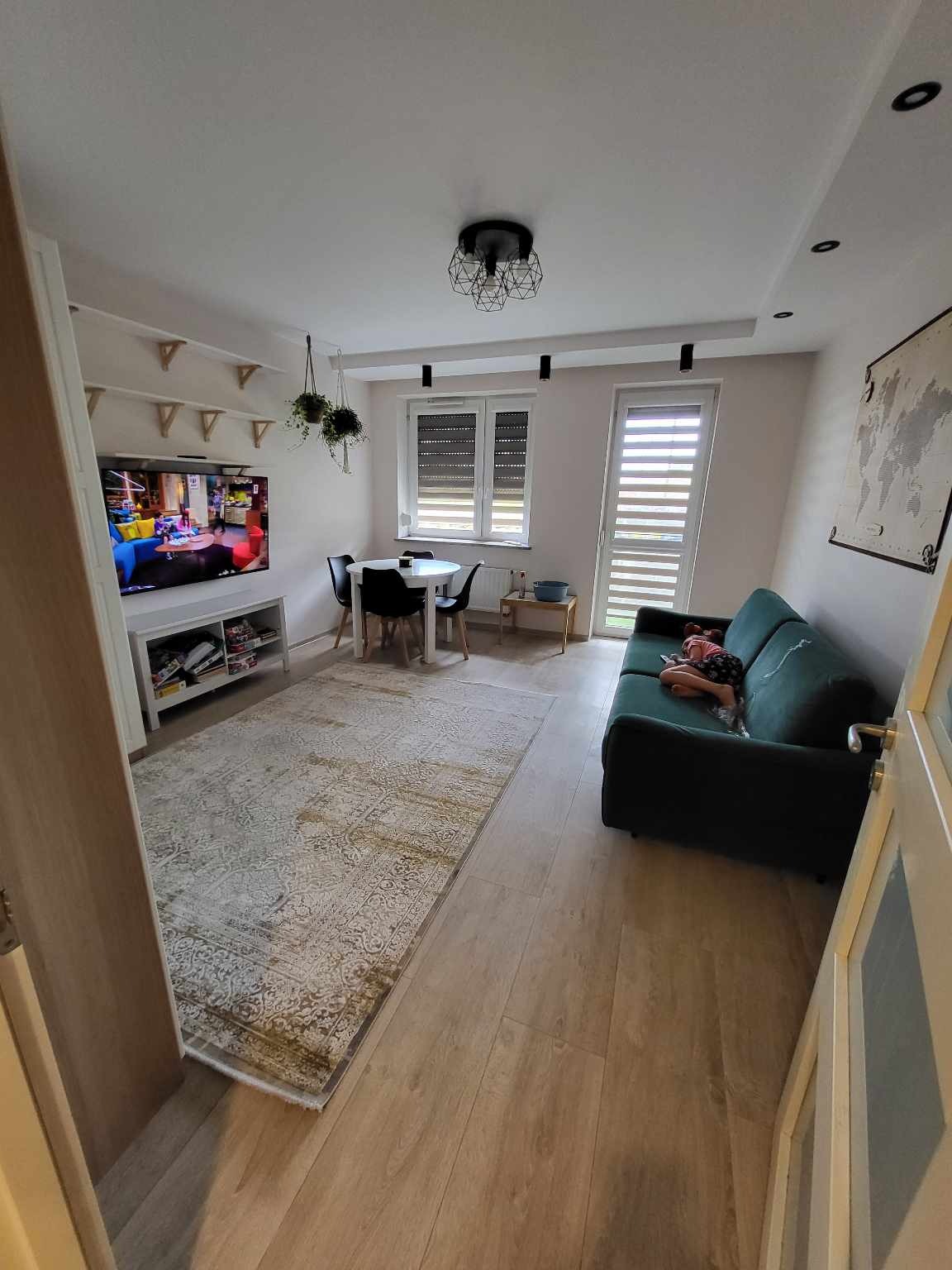 Jasne wnętrze pokoju dziennego z drewnianą podłogą, wzorzystym dywanem, zieloną sofą z leżącą osobą, stołem z krzesłami, telewizorem i dekoracjami ściennymi.