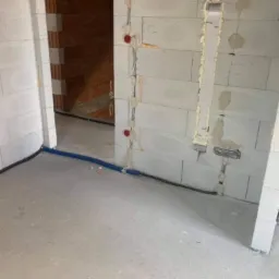 Wnętrze w trakcie prac wykończeniowych: widoczne kable elektryczne w peszlach prowadzone wzdłuż ściany z betonu komórkowego, puszki instalacyjne zamontowane w ścianie, pianka montażowa wokół...