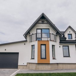 Safe Dom Investments - Nowoczesny, dwukondygnacyjny dom z białymi ścianami, ciemną stolarką okienną i grafitową bramą garażową, balkon z czarną balustradą, widok z poziomu trawnika.