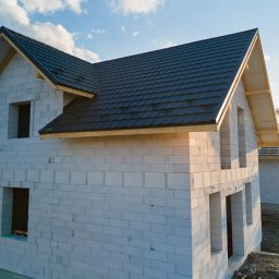 Safe Dom Investments - Surowy stan dwukondygnacyjnego domu z białego betonu komórkowego, z czarną, nową dachówką, widoczne otwory okienne i drzwiowe, w tle drugi dom w stanie surowym otwartym, błękitne niebo z chmurami.