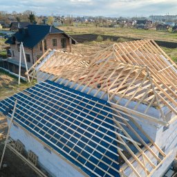 Safe Dom Investments - Wznoszenie drewnianej konstrukcji dachu na budynku z białych pustaków, z widocznym fragmentem niebieskiej membrany dachowej i drugim domem w tle, widok z lotu ptaka.