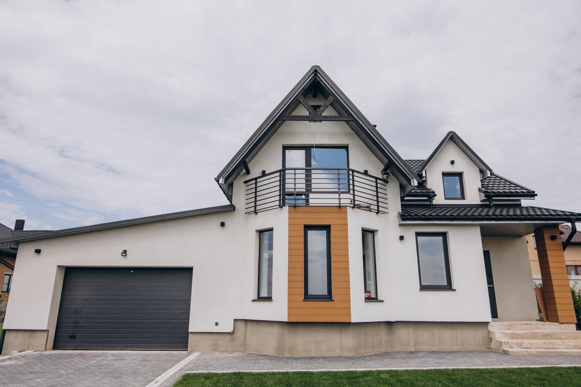 Nowoczesny, dwukondygnacyjny dom z białymi ścianami, ciemną stolarką okienną i grafitową bramą garażową, balkon z czarną balustradą, widok z poziomu trawnika.