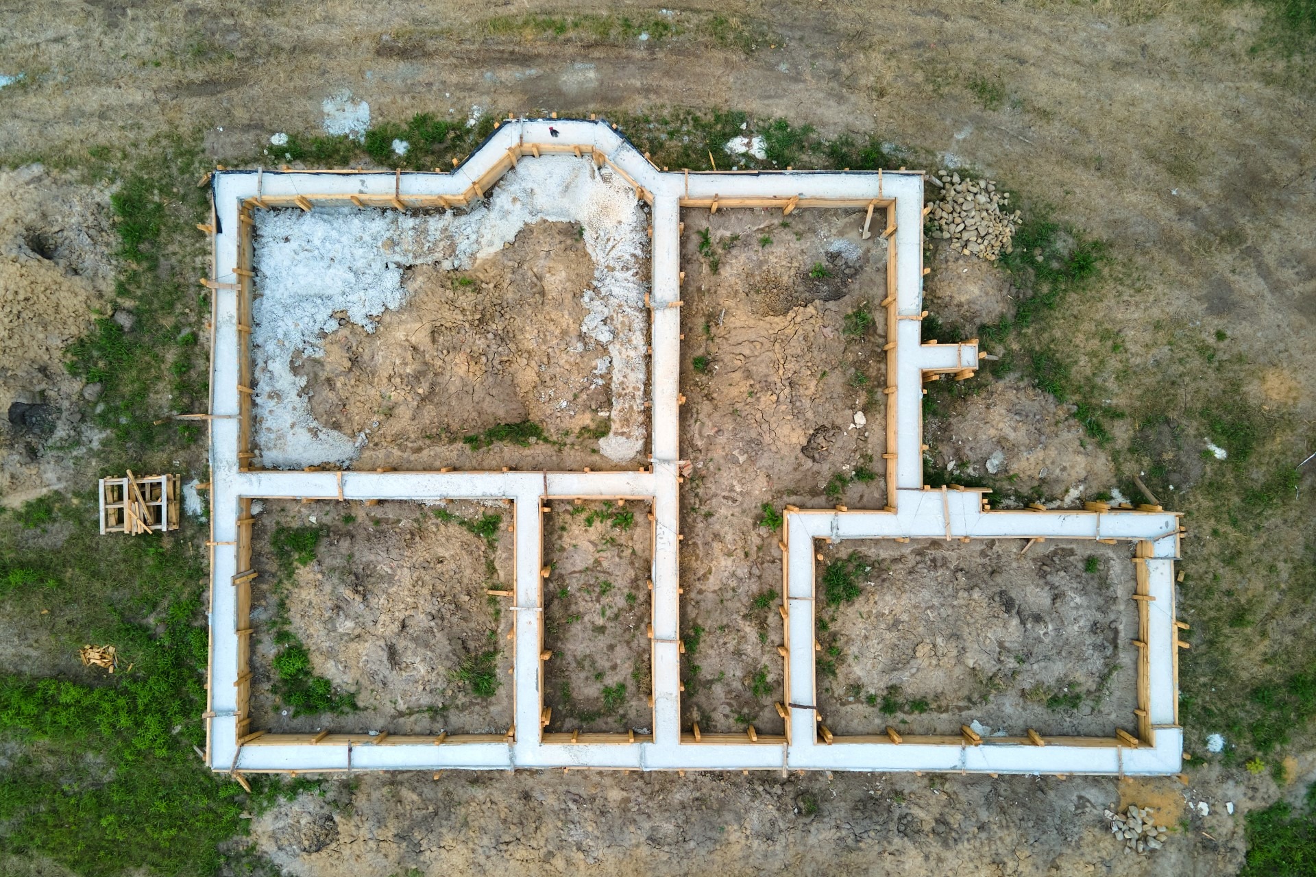 Widok z lotu ptaka na szalunek fundamentów domu jednorodzinnego na działce z widocznymi elementami drewnianymi i ubitą ziemią wewnątrz.