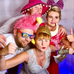 Grupa czterech osób w fotobudce, pozujących z rekwizytami: różowy kapelusz sombrero, okulary z napisem 'I SEE DRUNK PEOPLE', hełm wikinga i różowa boa z flaminga.