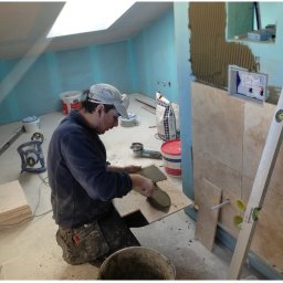 Kreative Home - Montaż beżowych płytek na ścianie w łazience, widoczny pracownik nakładający klej pacą na płytkę, poziomica oparta o ścianę, niebieskie ściany w tle, widoczny otwór okienny w dachu.
