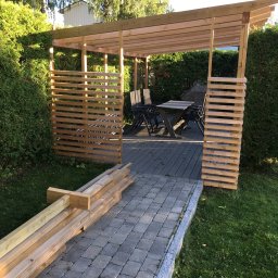 Kreative Home - Nowoczesna drewniana altana z ażurowymi ściankami, z meblami ogrodowymi na tarasie z desek kompozytowych, widok z kamiennej ścieżki, na trawie leżą przygotowane elementy konstrukcyjne.