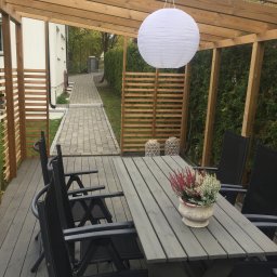 Kreative Home - Zadaszony taras z drewnianą pergolą, szarym stołem i krzesłami, dekorowany lampionem i donicą z wrzosami, widok na ścieżkę z kostki brukowej.