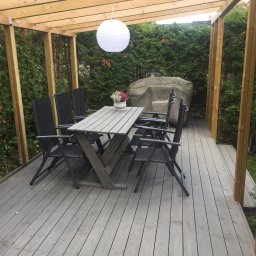 Kreative Home - Zadaszony taras z szarego drewna z drewnianą konstrukcją, stołem i krzesłami, dekorowany lampionem i roślinami w doniczce, z widocznym pokrowcem na grill marki Landmann.