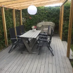 Zadaszony taras z szarego drewna z drewnianą konstrukcją, stołem i krzesłami, dekorowany lampionem i roślinami w doniczce, z widocznym pokrowcem na grill marki Landmann.