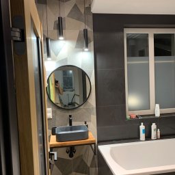 Kreative Home - Nowoczesna łazienka z umywalką nablatową w kształcie prostokąta, okrągłym lustrem i geometrycznymi płytkami na ścianie, oświetlona trzema cylindrycznymi lampami wiszącymi.