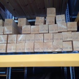 Kreative Home - Stos kwadratowych belek drewnianych z nadrukowanymi numerami ESAS ułożonych na żółtym regale w magazynie.