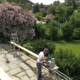 Kreative Home - Mężczyzna w szarej odzieży tnie deskę piłą elektryczną na zewnątrz, w tle bujna roślinność i drabina aluminiowa.