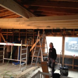 Kreative Home - Wnętrze domu w trakcie budowy, widoczne drewniane belki stropowe, ściany z izolacją, drabiny i rusztowania, oraz pracownik w ubraniu roboczym.