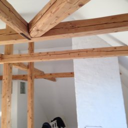 Kreative Home - Surowe drewniane belki stropowe w trakcie budowy, połączone metalowymi elementami, widoczne białe ściany i fragment okna dachowego, otwarte drzwi w tle.