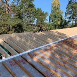 Kreative Home - Konstrukcja dachu domu w trakcie budowy, widoczne krokwie i membrana dachowa z nadrukiem, na tle zielonych drzew i błękitnego nieba.