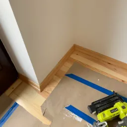 Montaż drewnianej listwy przypodłogowej z użyciem gwoździarki Ryobi Air-Strike, zabezpieczone kartonem i niebieską taśmą malarską, widoczny fragment okna i gniazdko elektryczne