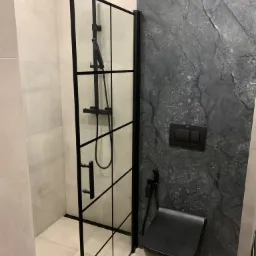 Nowoczesna łazienka z czarną kabiną prysznicową typu walk-in z profilami, czarną toaletą wiszącą i ścianą wykończoną płytami imitującymi ciemny marmur.