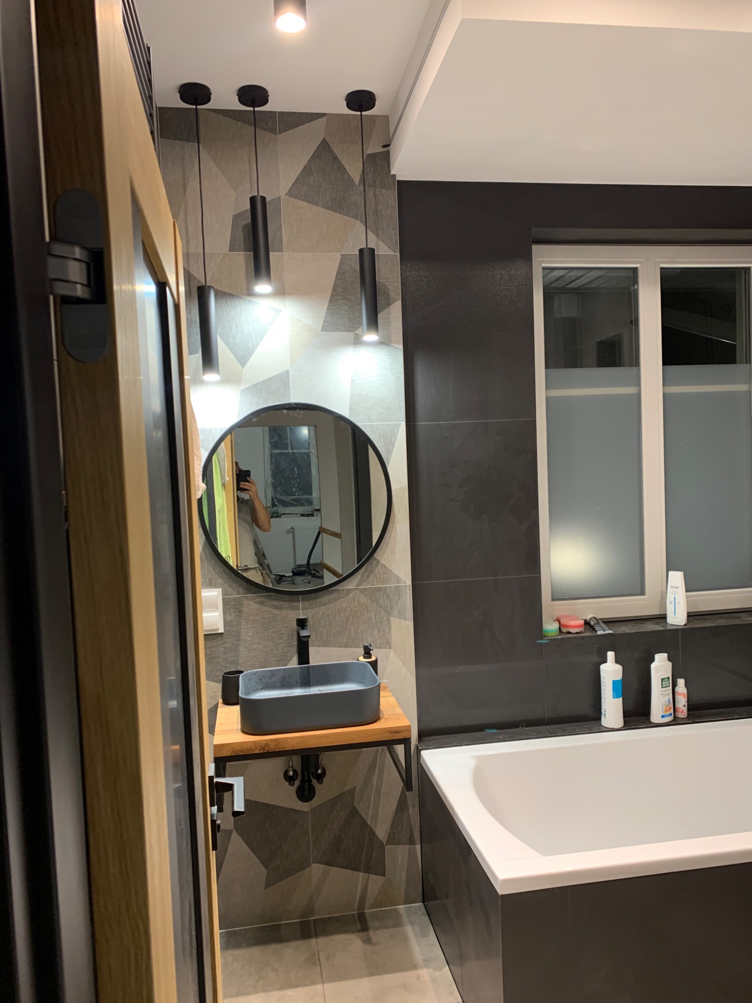 Nowoczesna łazienka z umywalką nablatową w kształcie prostokąta, okrągłym lustrem i geometrycznymi płytkami na ścianie, oświetlona trzema cylindrycznymi lampami wiszącymi.