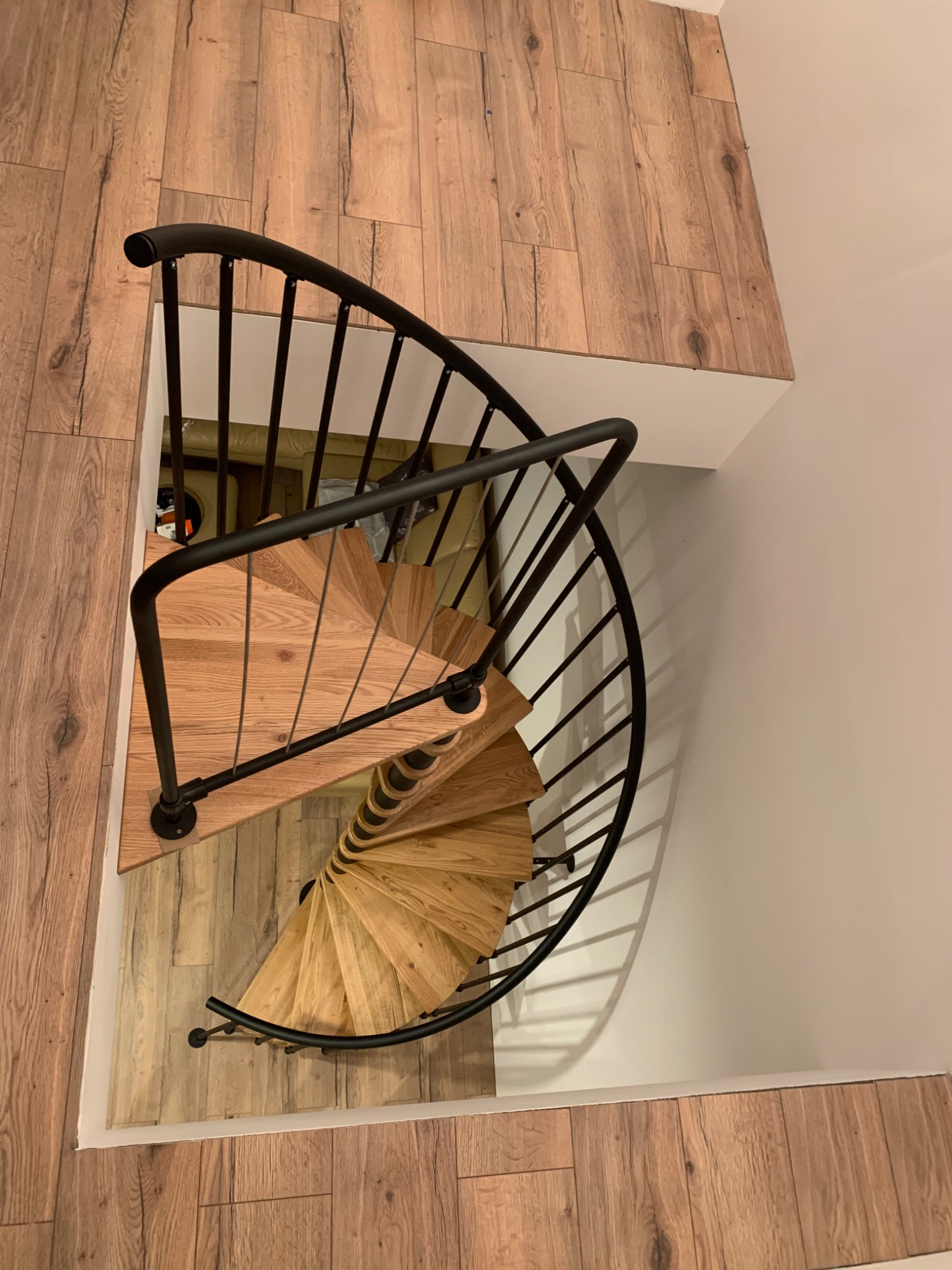 Widok z góry na drewniane schody spiralne z czarną metalową balustradą, otoczone panelami podłogowymi imitującymi drewno i białą ścianą.