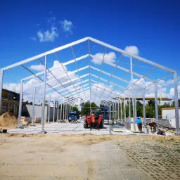 Montaż stalowej konstrukcji hali na betonowej płycie, widoczny podnośnik i pracownicy, w tle budynki i błękitne niebo z chmurami.