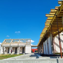 Forma Budownictwo Paweł Dąbrowski - Wznoszenie budynku z białych bloczków, widoczne szalunki stropowe wsparte na stalowych podporach, w tle błękitne niebo i częściowo ukończony dach z drewnianą konstrukcją.