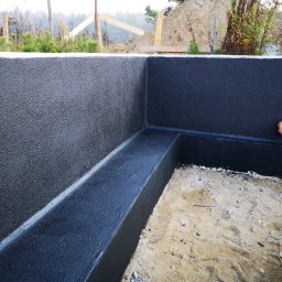 Forma Budownictwo Paweł Dąbrowski - Zabezpieczone hydroizolacją fundamenty domu w trakcie budowy, widoczna warstwa izolacyjna w kolorze czarnym oraz otwór kanalizacyjny zabezpieczony niebieską folią.