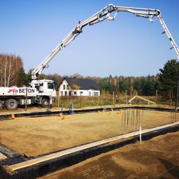 Forma Budownictwo Paweł Dąbrowski - Wylewanie betonu na fundamenty domu jednorodzinnego za pomocą pompy do betonu KB Beton. Widoczne zbrojenia, szalunki i izolacja pionowa fundamentów. W tle dom w stanie surowym otwartym.