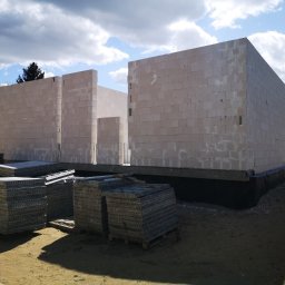 Forma Budownictwo Paweł Dąbrowski - Wznoszone ściany parterowego budynku z bloczków z betonu komórkowego na fundamencie z izolacją przeciwwilgociową, widoczne ułożone palety z bloczkami na piaszczystym podłożu, pochmurne niebo w tle.