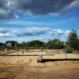 Przygotowany teren pod fundamenty domu jednorodzinnego z widocznymi zbrojeniami, szalunkami i piaskiem zasypowym na tle błękitnego nieba z chmurami i zabudowy jednorodzinnej.