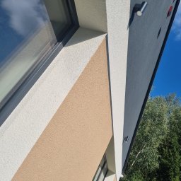 Forma Budownictwo Paweł Dąbrowski - Fragment elewacji budynku z widocznym oknem, oświetleniem zewnętrznym i fragmentem zieleni w tle; elewacja w odcieniach beżu, bieli i szarości.